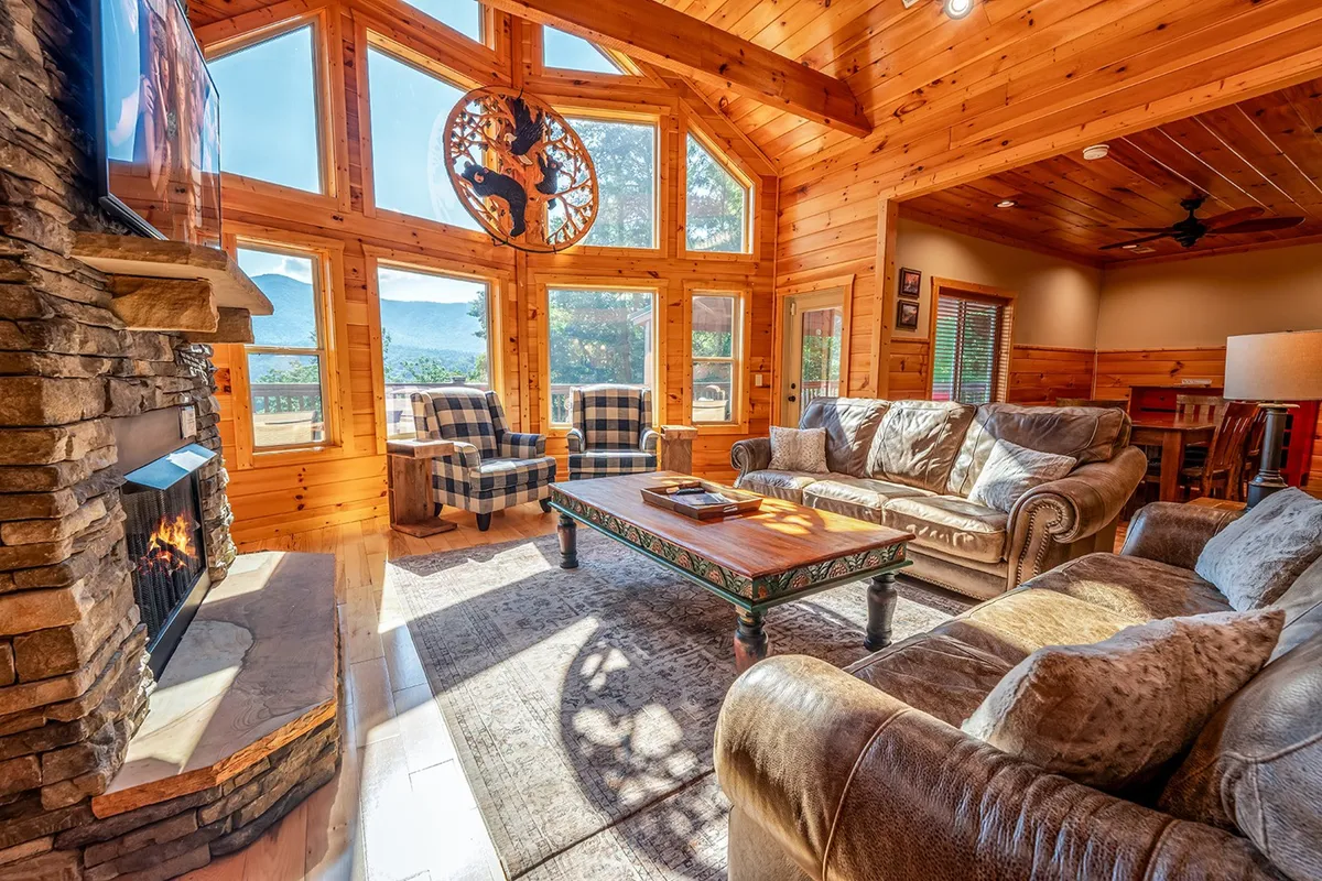 An inviting living area with large windows offering panoramic mountain views, a comfortable sofa, a coffee table, a glowing fireplace, and classic decorative accents. The fire and the mountain backdrop create a tranquil and scenic ambiance.