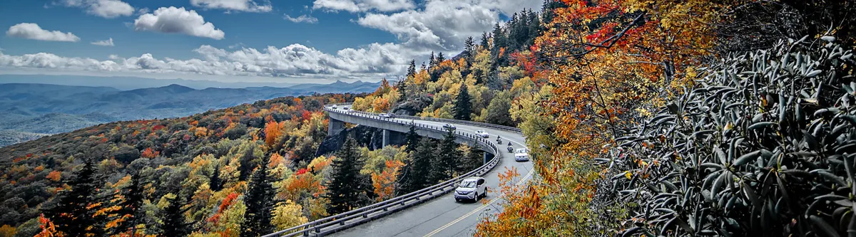 This image captures a curving road winding through vibrant autumn foliage, with distant mountains and a cloudy sky stretching overhead. The scene conveys the natural beauty and sense of adventure found just outside Bearmont Lodge, inviting families to envision themselves exploring the surrounding Great Smoky Mountains National Park.