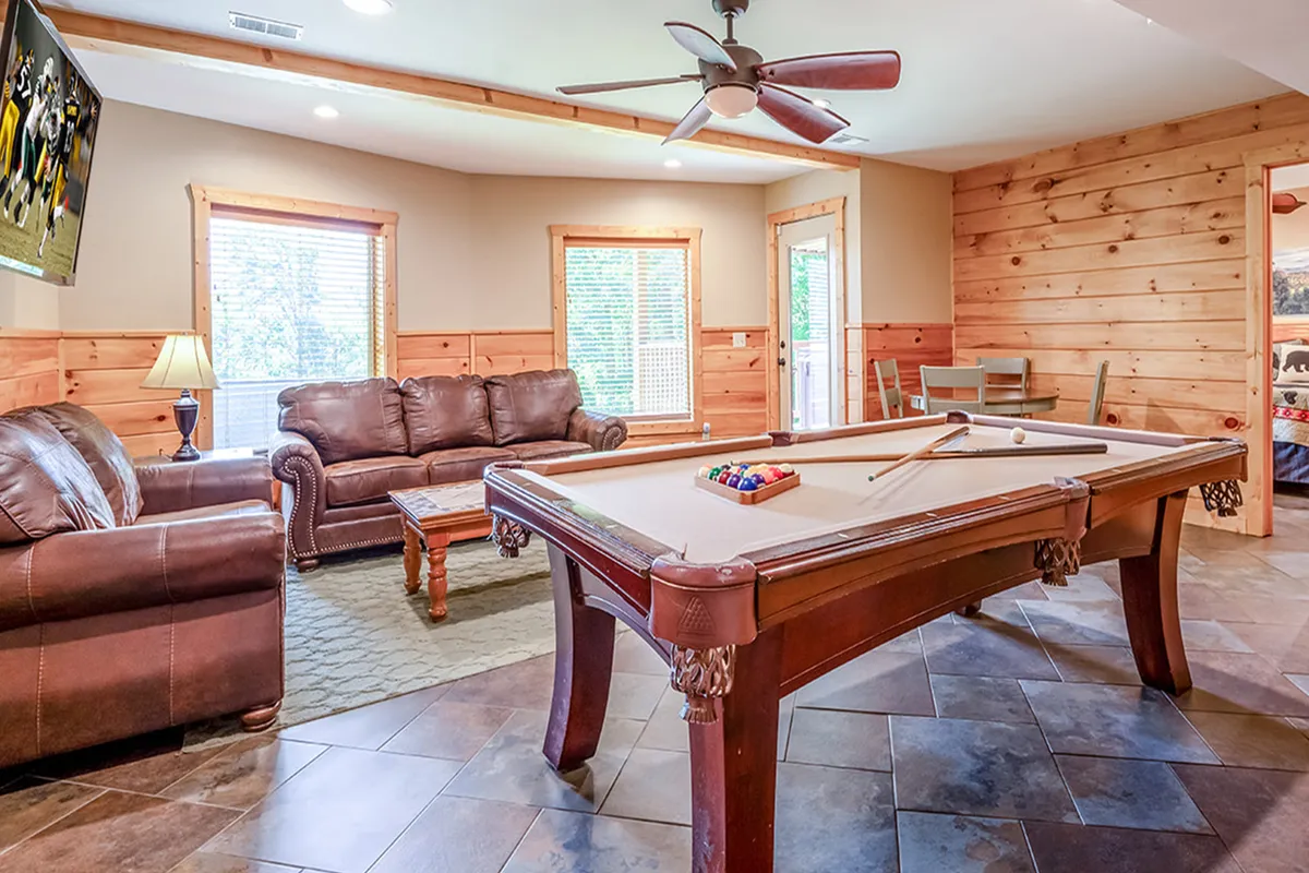 This image features a spacious game room with a pool table in the center, surrounded by leather sofas for comfortable seating. Large windows allow plenty of natural light, and a TV is mounted on the wall for watching sports or movies. The arrangement invites socializing and relaxation, with clear views of the major amenities.