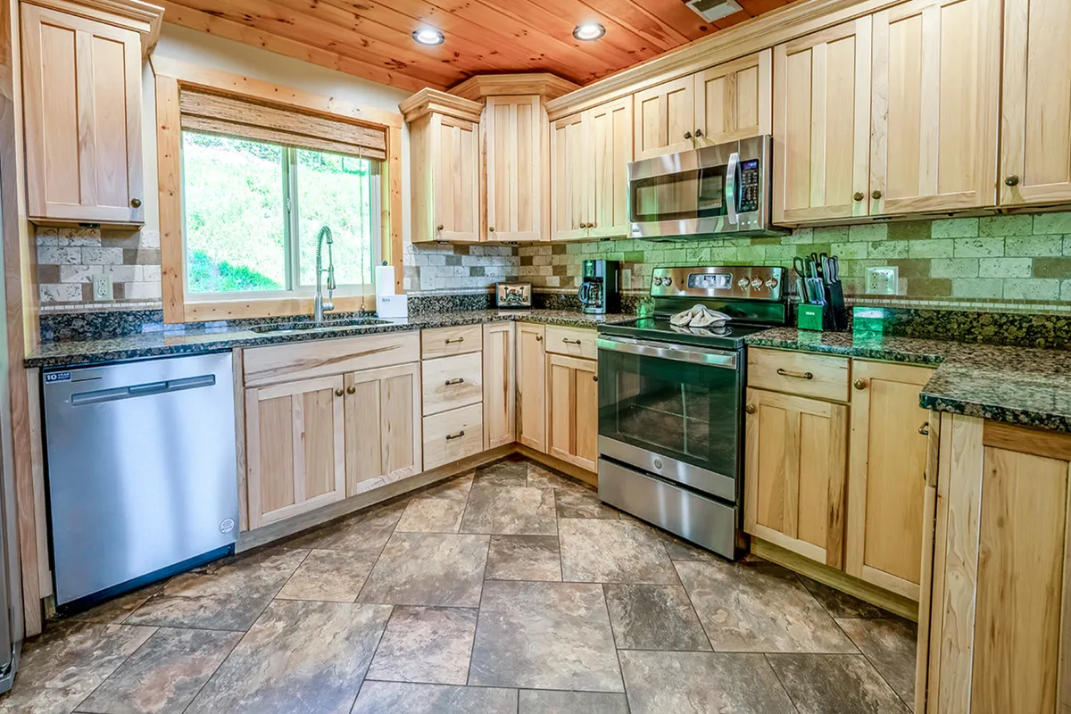 Another angle of a kitchen with granite countertops, wooden cabinets, stainless steel appliances, and a large window allowing natural light to fill the space. The functional design supports meal prep for groups or families.