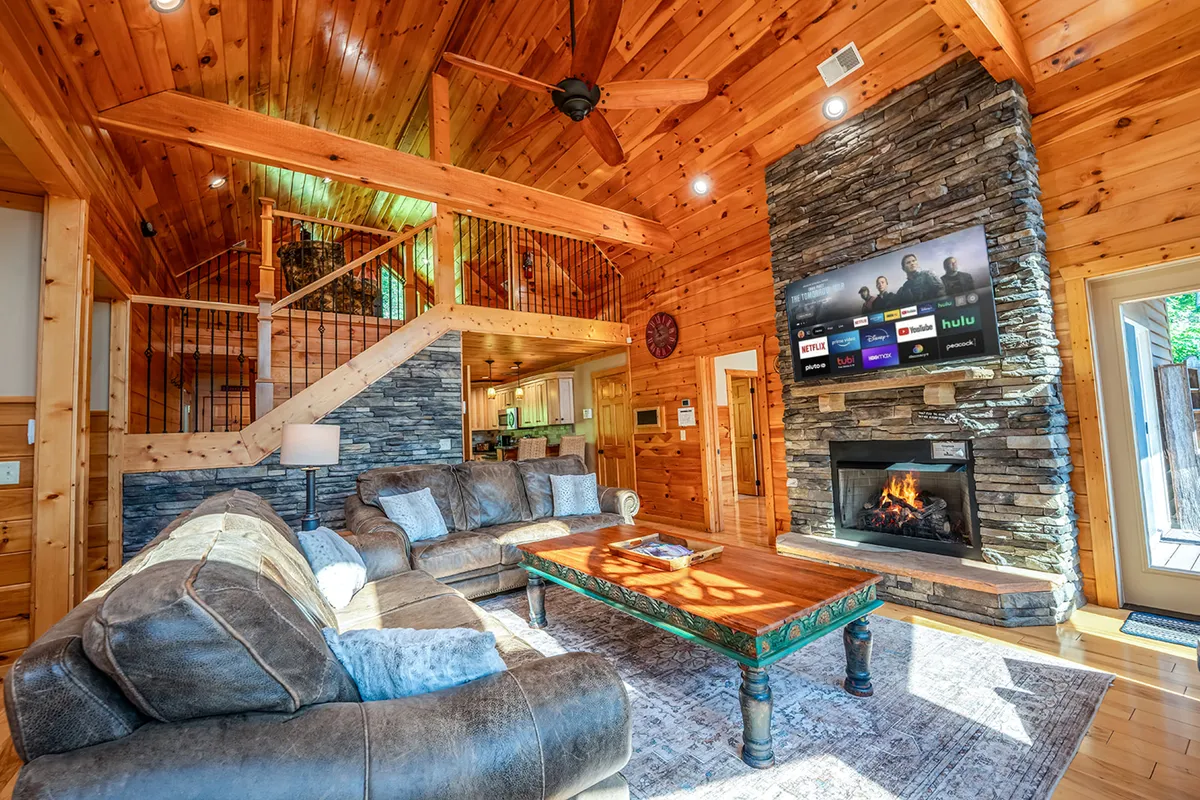 A spacious living room featuring a prominent stone fireplace, a large sectional sofa arranged for gatherings, a wooden coffee table, and a TV with streaming services. Natural light pours in, highlighting the cozy and welcoming atmosphere ideal for family activities.
