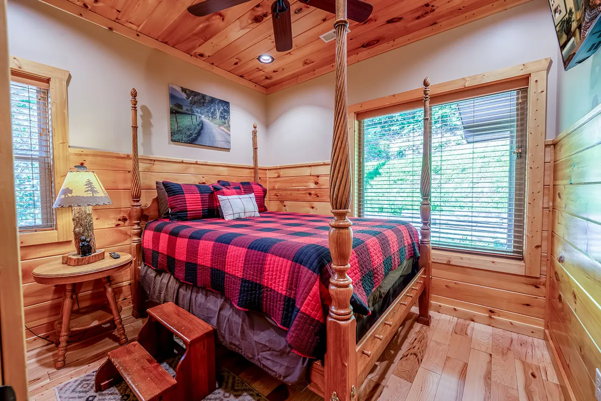 A warm and inviting bedroom featuring a four-poster bed with plaid bedding, wooden furniture, a nature-inspired lamp, and framed artwork. The space exudes comfort and a rustic charm, ideal for restful nights after a day of adventure.