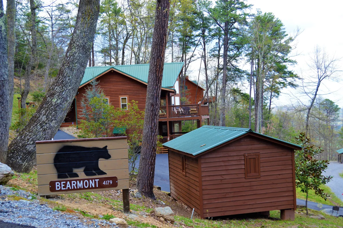 Rustic wooden cabins nestled among tall trees, accompanied by a playful bear sign and surrounded by the lush forest. The cabins evoke a sense of adventure and immersion in nature, hinting at nearby trails and wildlife encounters.