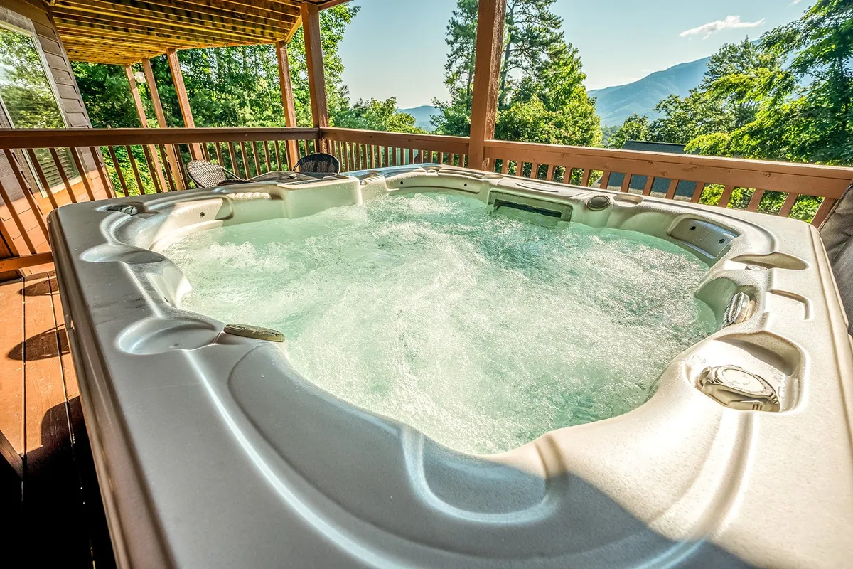 A serene outdoor hot tub sits on a deck surrounded by lush greenery, with stunning mountain views in the distance. The spacious setting invites relaxation and features seating for several guests, ideal for unwinding after a day of adventure.
