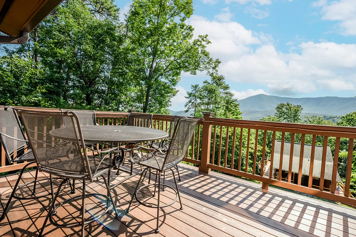 An outdoor dining table sits on a deck shaded by surrounding trees, with a panoramic view of the mountains in the background. The tranquil setting is perfect for family gatherings, al fresco meals, and soaking up the natural scenery from the comfort of home.