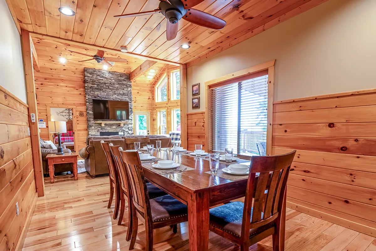 A warm dining area set with a family-friendly dining table and chairs, a matching stone fireplace, and large windows letting in natural light. The layout encourages shared meals, celebrations, and relaxed gatherings by the fire.