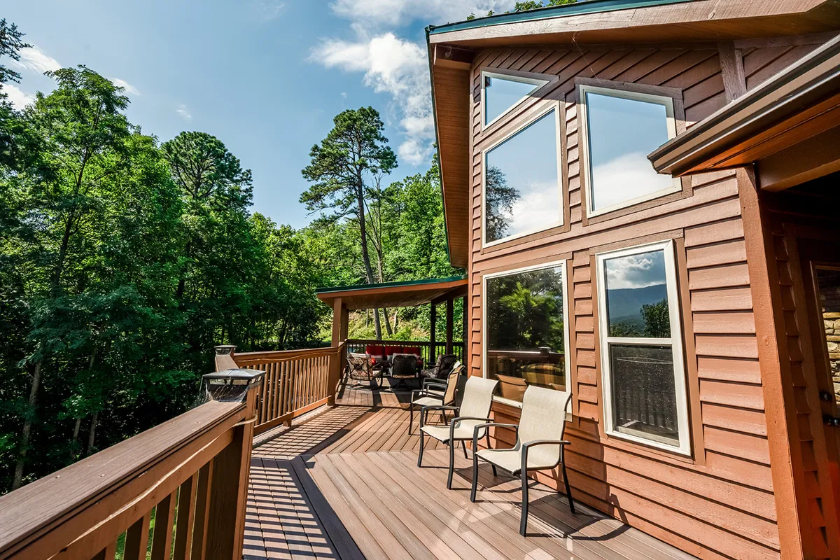 A spacious wooden deck with comfortable outdoor seating, surrounded by lush trees. The deck features large windows that reflect the forested landscape, and the area is set up for relaxing and socializing with friends. The serene backdrop and open-air design make it ideal for group gatherings.